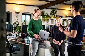 Zwei e-pixler-Mitarbeiterinnen im Gespräch, eine hält einen Laptop, während ein Kollege spricht.