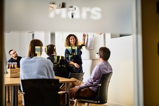 e-pixler-Mitarbeiter in einem Meetingraum, eine Kollegin präsentiert Ideen an einem Whiteboard.