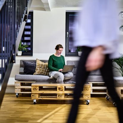 Mitarbeiterin bei e-pixler arbeitet mit einem Laptop auf einer Couch aus Paletten in einem modernen Büro mit Pflanzen und Holzfußboden.