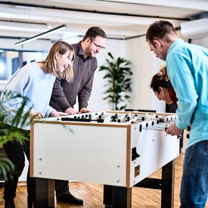 e-pixler-Mitarbeiter spielen Tischkicker in einem modernen Empfangsbereich mit Pflanzen und Holzboden.