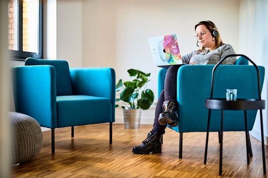 e-pixler-Mitarbeiterin arbeitet mit Kopfhörern und Laptop in einem Lounge-Bereich mit blauen Sesseln und Pflanzen.