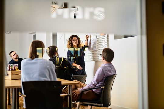 e-pixler-Mitarbeiter in einem Meetingraum, eine Kollegin präsentiert Ideen an einem Whiteboard.