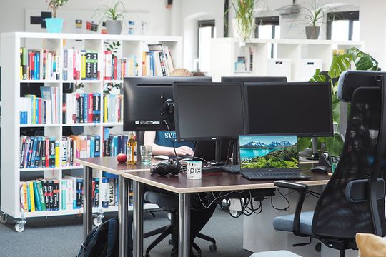 Arbeitsbereich bei e-pixler mit mehreren Monitoren und Regalen voller Bücher.