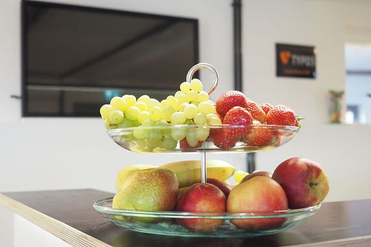 Obstschale mit Trauben, Äpfeln und Bananen auf einem Tisch im Büro von e-pixler, im Hintergrund ein Fernseher.